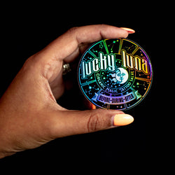 Hand holding a colorful iridescent holographic circle sticker with 'Lucky Luna' branding  black print on rainbow background.  Text reads. lucky luna organic magic tatto salve before during after.