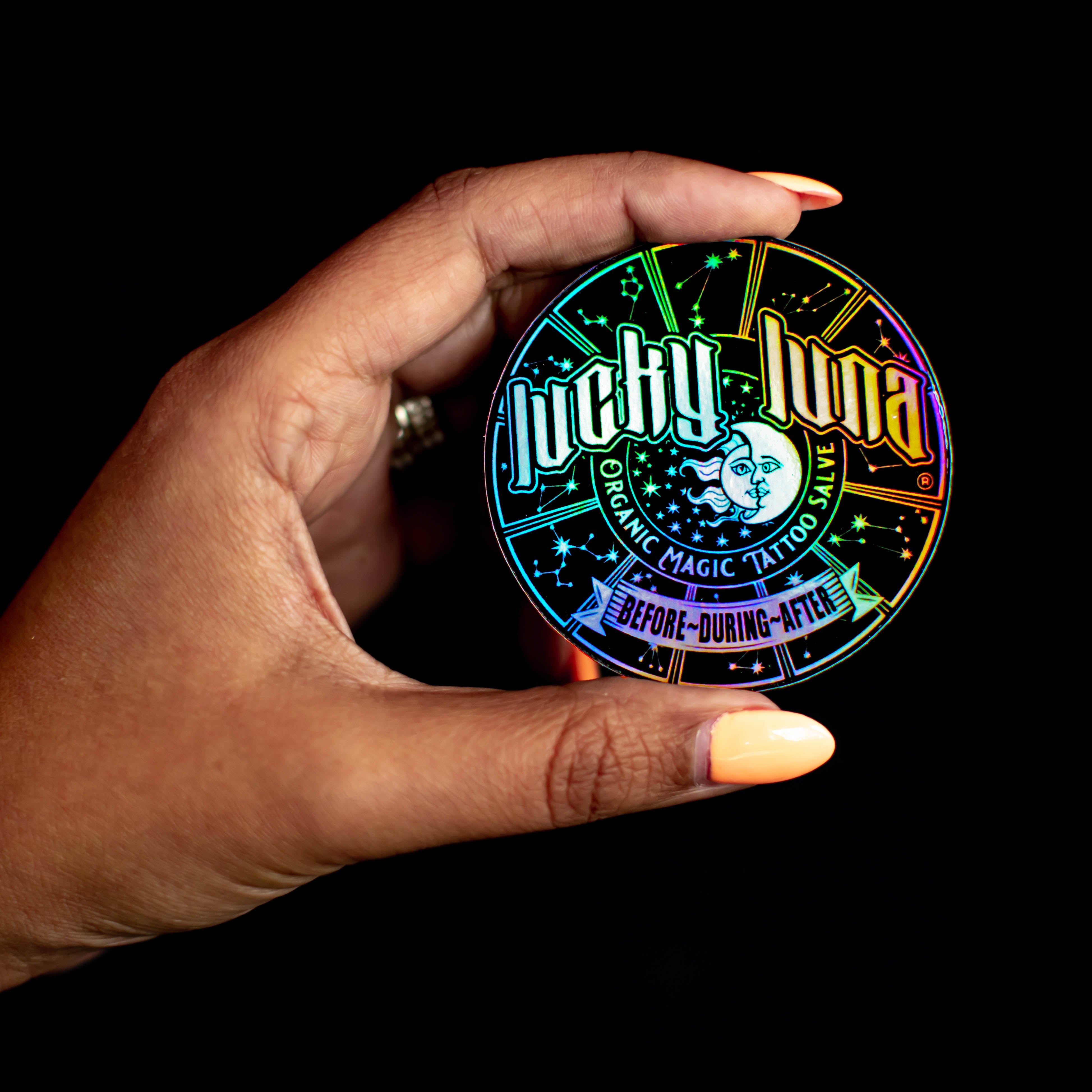 Hand holding a colorful iridescent holographic circle sticker with 'Lucky Luna' branding  black print on rainbow background.  Text reads. lucky luna organic magic tatto salve before during after.